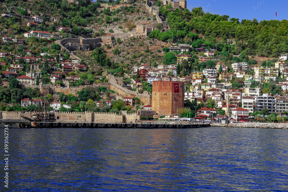 Alanya coastline with modern cottages, an ancient wall and Red Tower on the side of a mountain against a backdrop of blue water. Sunny summer panorama of Turkish town - view from the Mediterranean Sea