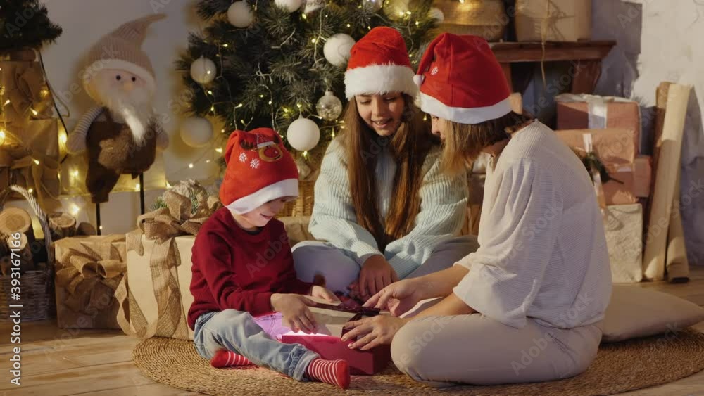 Mom with children in Santa hats sitting on the carpet near the Christmas tree and decorations open a gift with light inside Mom with children in Santa hats sitting on the carpet near the Christmas tree and decorations open a gift with light inside