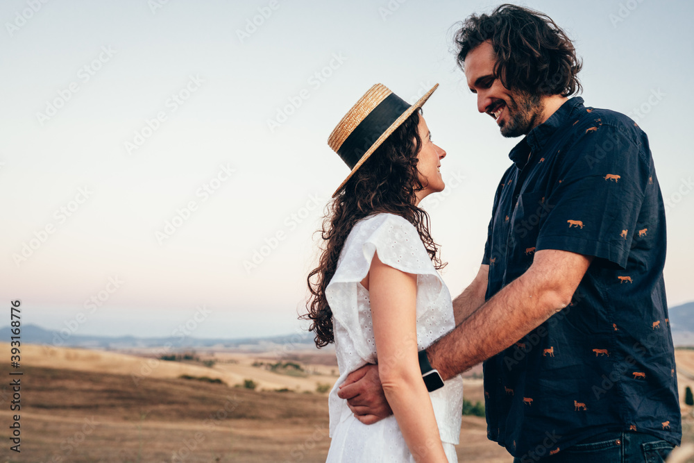 a boy and a girl in love hug each other as they look into each other's eyes in the middle of the summer countryside at sunset