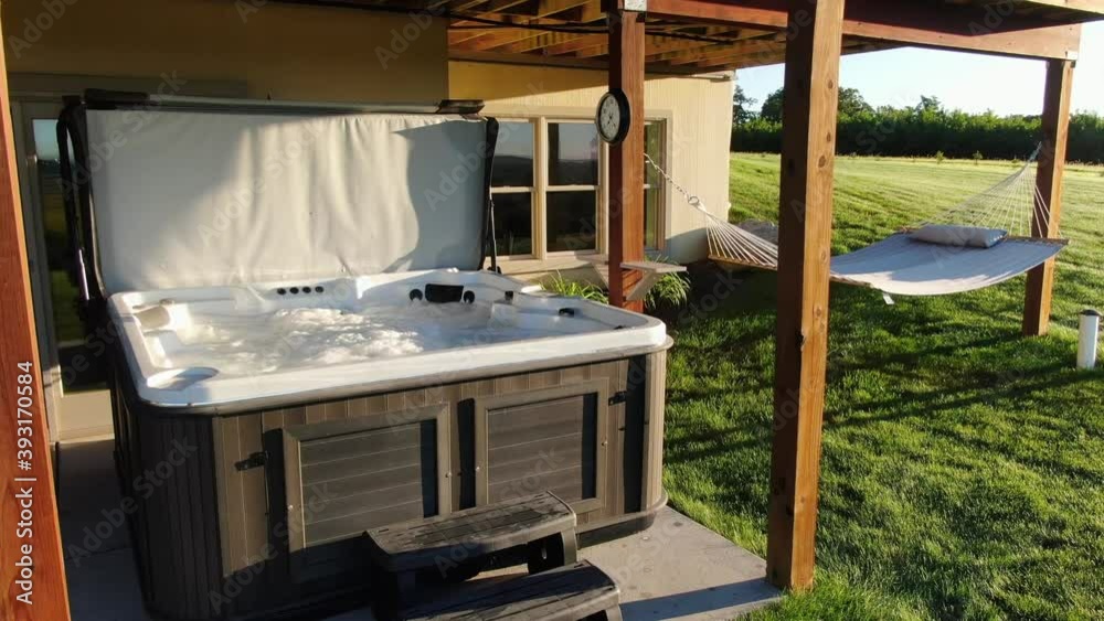 Outdoor hot tub, open and ready for therapy treatment. Water bubbles and steams in morning light. Hammock nearby. Vacation, relaxation getaway theme.