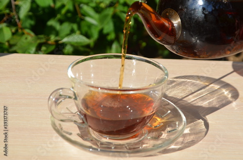 A girl pours tea from a transparent teapot into a transparent cup against a background of green foliage Good morning, outdoor cafe, energy boost. Tea being poured into glass tea