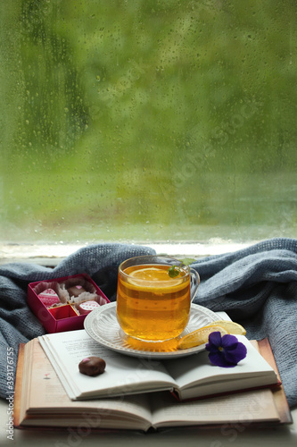 A cup of tea with lemon, books and candies on the background of a window on a rainy day.
