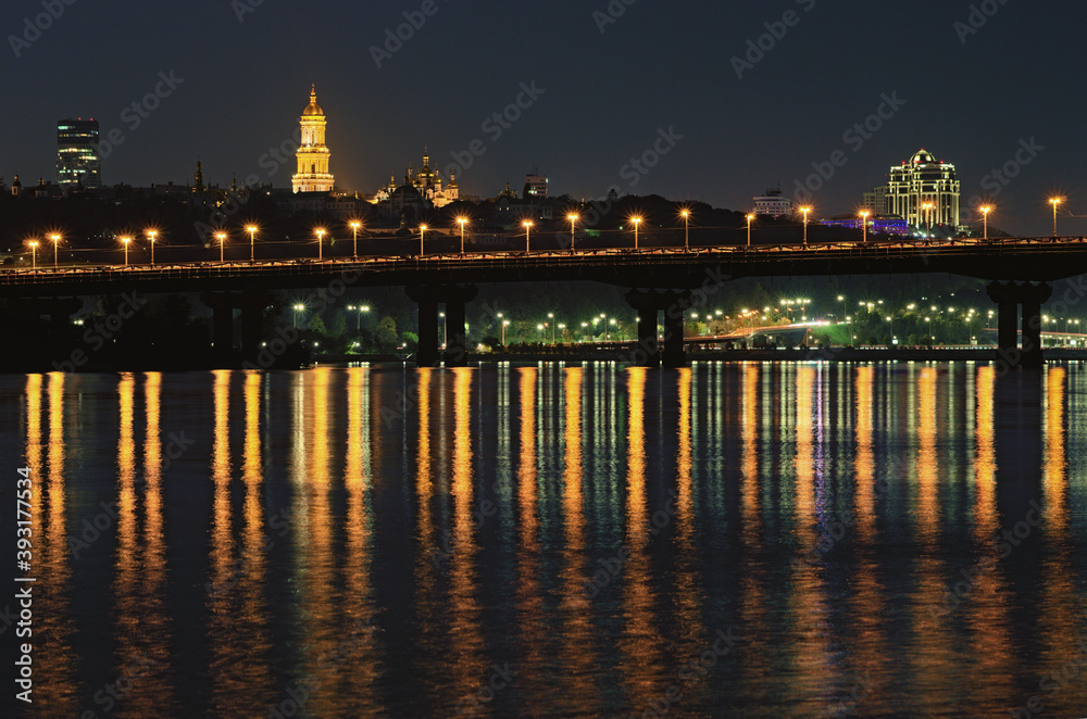 Night cityscape with Paton bridge over Dnieper river. Light from lanterns reflected in the water. Illuminated Bell tower of Kyiv Pechersk Lavra at the background. City lights reflected in the water