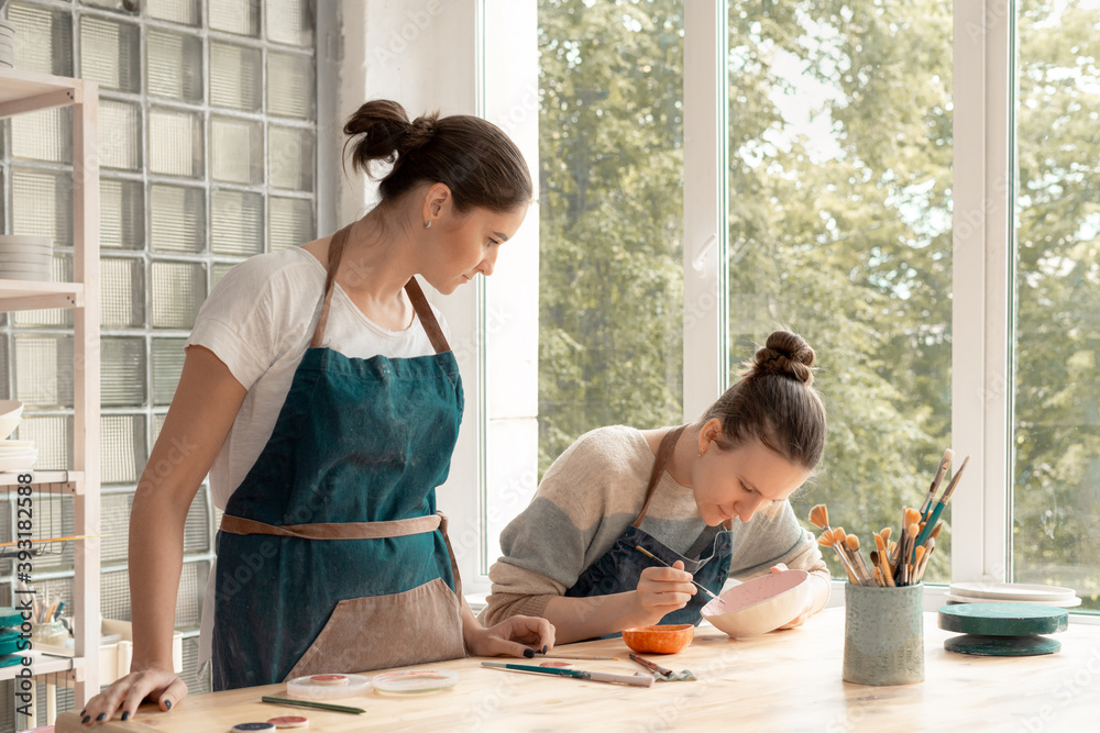 Woman making ceramic pottery. Attractive skilled young lady in apron standing at table and teaching student. Concept for workshop and master class Woman making ceramic pottery. Attractive skilled young lady in apron standing at table and teaching student. Concept for workshop and master class