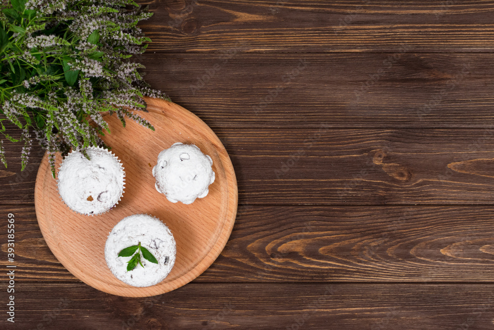 Cupcakes on a background of flowers and mint leaves, on a round board on a wooden background. Place for gadpis. Cupcakes on a background of flowers and mint leaves, on a round board on a wooden background. Place for gadpis.
