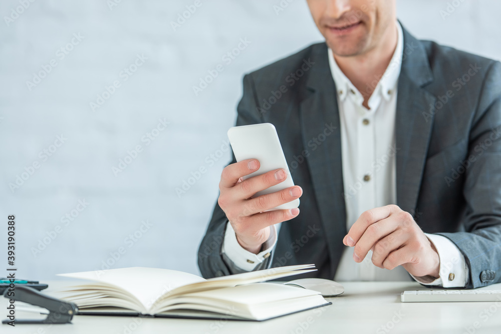 Cropped view of positive businessman using smartphone, while sitting at table on blurred background Cropped view of positive businessman using smartphone, while sitting at table on blurred background