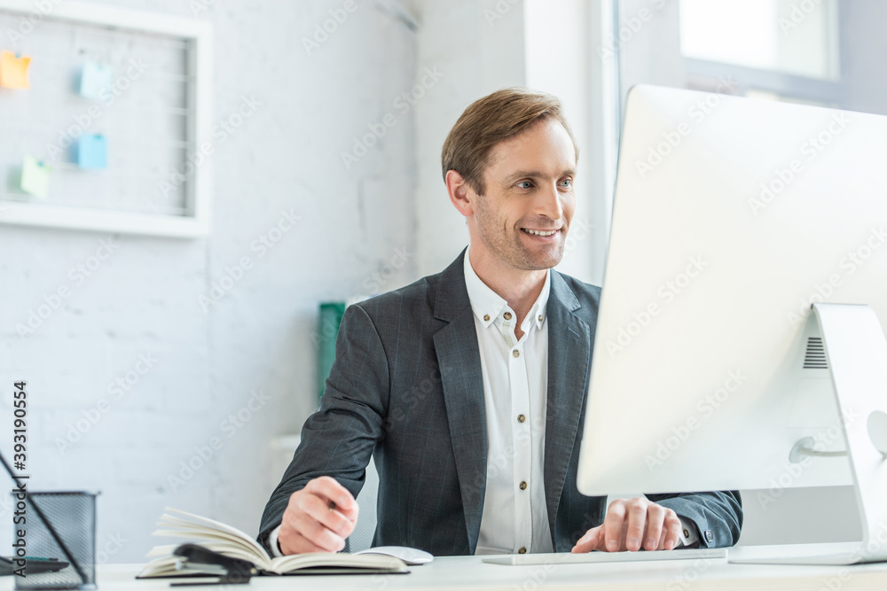 Happy businessman looking at computer monitor, while sitting at workplace in office on blurred background