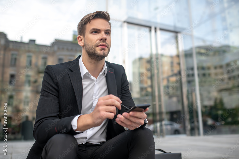 A businessman writing a message on a smartphone and looking thoughtful A businessman writing a message on a smartphone and looking thoughtful