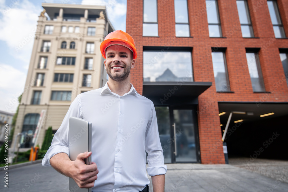 Male engineer in protective helmet standing near the building and looking positive