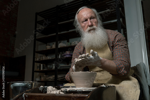 Old male potter. High angle of craftsman working on pottery wheel while sculpting from clay pot in workshop. Focus on arms. Concept of ceramic art