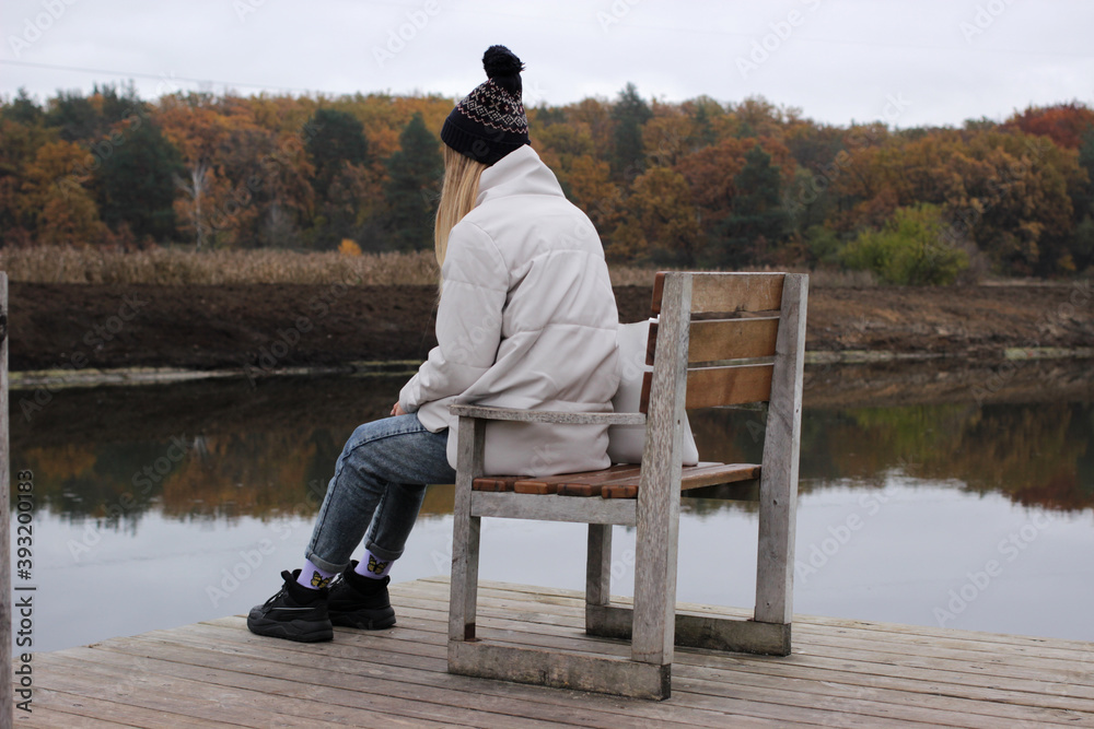 A young woman, warmly dressed, in a winter white jacket and hat, sits alone on a chair on a wooden pier near the river bank. View from the back. A young woman, warmly dressed, in a winter white jacket and hat, sits alone on a chair on a wooden pier near the river bank. View from the back.