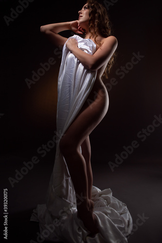 a naked beautiful woman covers herself with a white sheet on a dark background