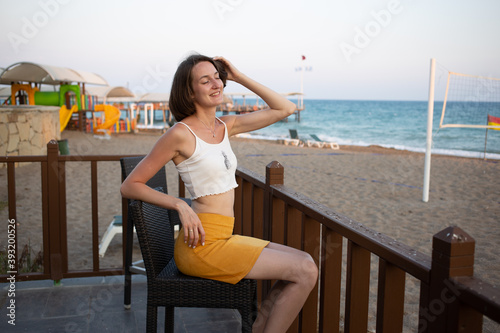 A girl in a yellow skirt and white top sits at a chair on the summer terrace of a cafe. On vacation in Turkey. Beach and sea in the background. Smile. Sunny day. Vocation. Lifestyle. Rest. Relax.