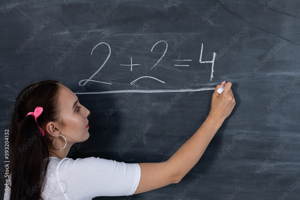 Teenager chalks a math equation on a dark gray school blackboard. Her hair is pinned with a pink ribbon in two ponytails.
