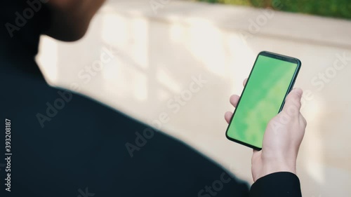 Over the shoulder shot of man using a phone with a green screen outdoors. Handsome man scrolling through smartphone apps outside in the city.