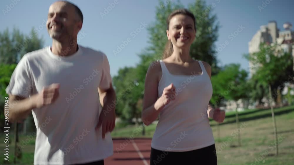 Portrait of smiling Caucasian adult man and woman jogging on running track outdoors on sunny summer day. Positive sportsman and sportswoman training on sports field. Healthy lifestyle. Portrait of smiling Caucasian adult man and woman jogging on running track outdoors on sunny summer day. Positive sportsman and sportswoman training on sports field. Healthy lifestyle.