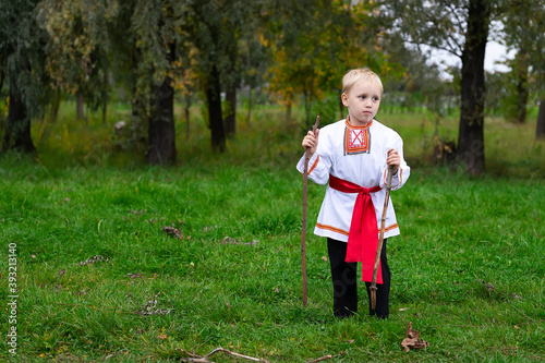 a boy in a Russian national costume stands in the grass in his hands two wooden sticks