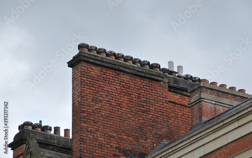 A large row of old fashioned traditional clay chimney pots on a red brick support against a grey sky from when buildings where built with coal burning heating