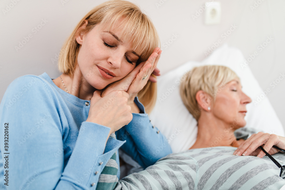 Beautiful middle age woman spending some time with her mother in hospital or nursing center. Senior woman lying in bed.