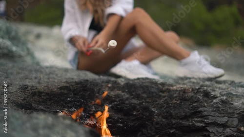 Slim young woman with sporty legs holds long wooden stick with marshmallow in hands and roasts over unique Turkish mountain fire sitting on rock closeup slow motion.