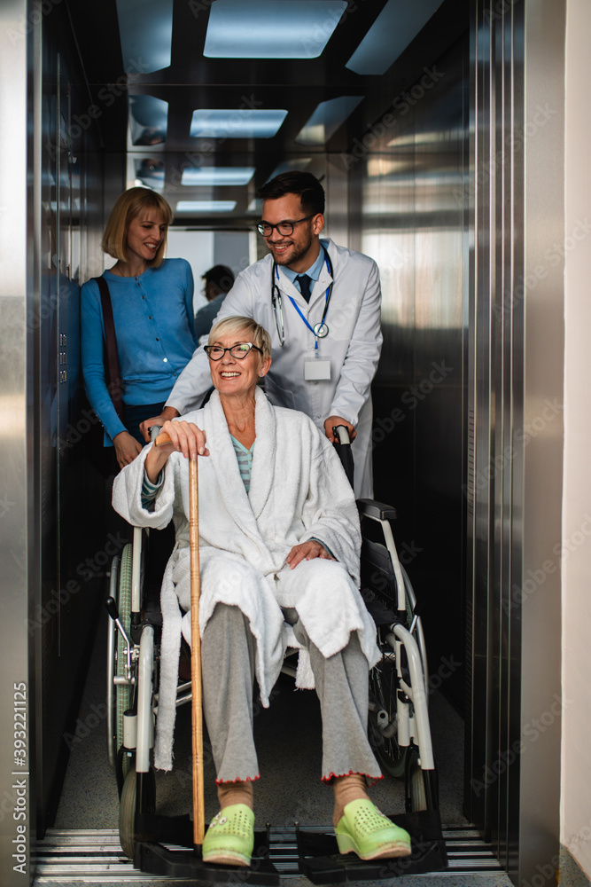 Young male practitioner talking with middle age woman and her sick mother which sitting in disability chair. They are standing in hospital or nursing center elevator. Young male practitioner talking with middle age woman and her sick mother which sitting in disability chair. They are standing in hospital or nursing center elevator.