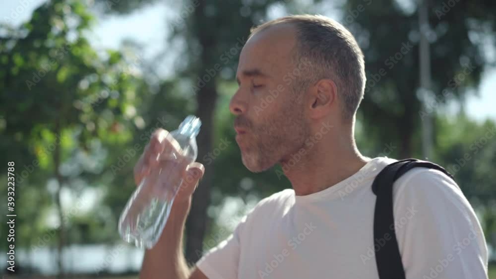 Close-up side view of handsome adult Caucasian man drinking water in sunlight outdoors and smiling at camera. Portrait of confident fit sportsman refreshing after workout or training. Close-up side view of handsome adult Caucasian man drinking water in sunlight outdoors and smiling at camera. Portrait of confident fit sportsman refreshing after workout or training.