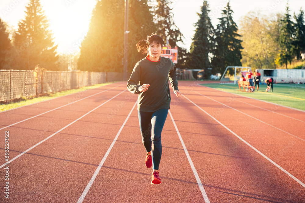 Asian woman running on the track in the winter. Sun flare suggests sunrise or sunset.