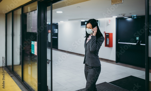 Attractive middle age business woman with protective face mask in modern business building. She is positive and using her smart phone. Coronavirus or Covid-19 pandemic lifestyle concept.