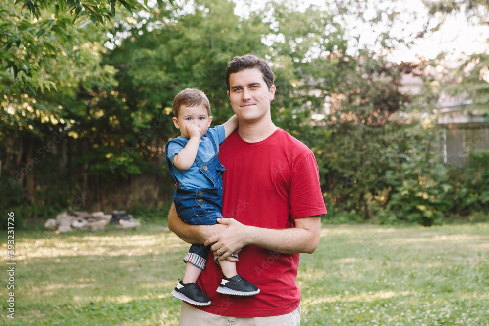 Fathers Day. Happy Caucasian father holding carrying baby boy on hands at backyard. Parent with child son outdoors. Authentic lifestyle touching tender moment. Single dad family life. Fathers Day. Happy Caucasian father holding carrying baby boy on hands at backyard. Parent with child son outdoors. Authentic lifestyle touching tender moment. Single dad family life.