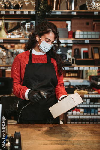 Beautiful and positive female cashier with face protective mask working on cash register in a modern supermarket or grocery store. Pandemic or epidemic lifestyle and consumerism concept.
