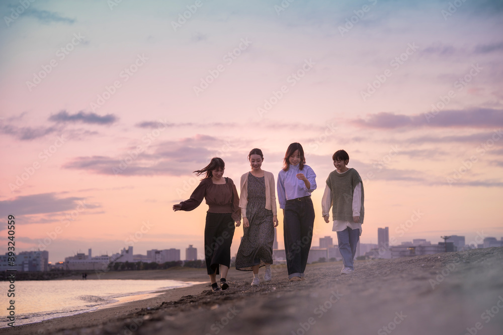夕暮れの中、寄り添って海沿いを歩く女性4人