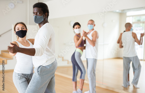Young Hispanic woman and African American man in protective face masks learning to dance waltz during group training. New life reality in pandemic