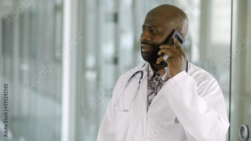 An African male doctor received an upsetting phone call. The man is upset and sad at receiving the new as he talks on the phone in the hospital corridor. He is wearing a lab coat and stethoscope.
