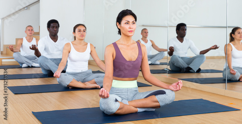 Focused asian woman sitting on mat in fitness center, making yoga meditation in lotus pose with group of people.