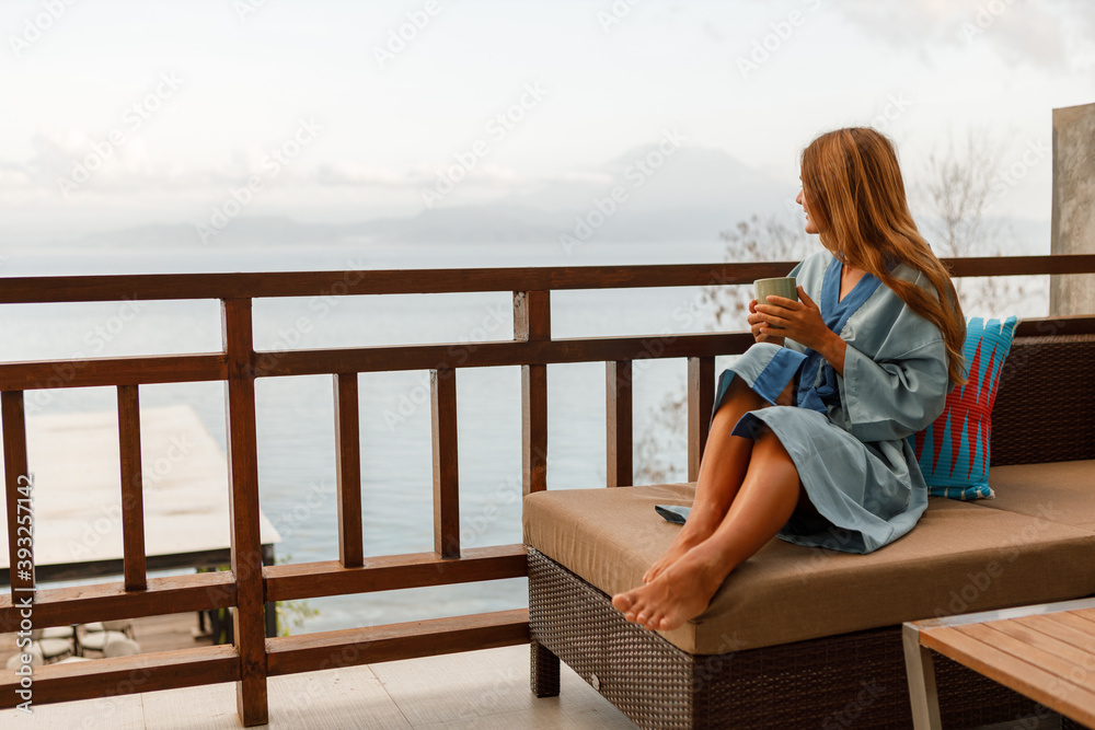 Young woman in bathrobe drinking coffee on balcony in the morning, look at the sea. Vacation on tropical island