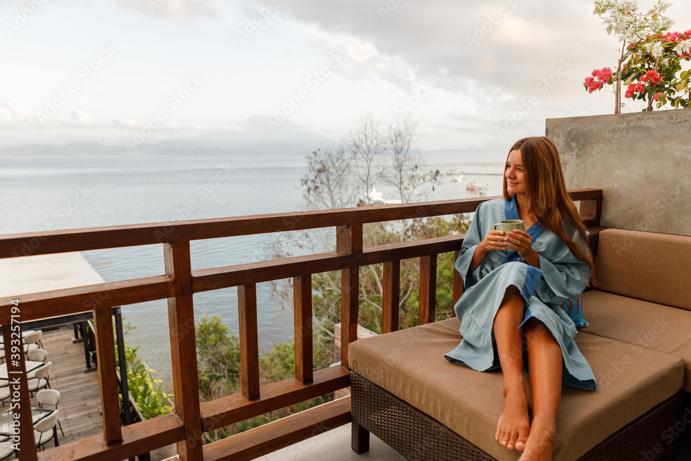 Young woman getting rest in bathrobe on terrace and drink coffee, wearing blue bathrobe and look at the sea
