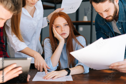 Close-up of exhausted overworked young business woman under stress at office. Colleagues shaking paper documents on blurry background. Business team wants to get answer and decision from boss.