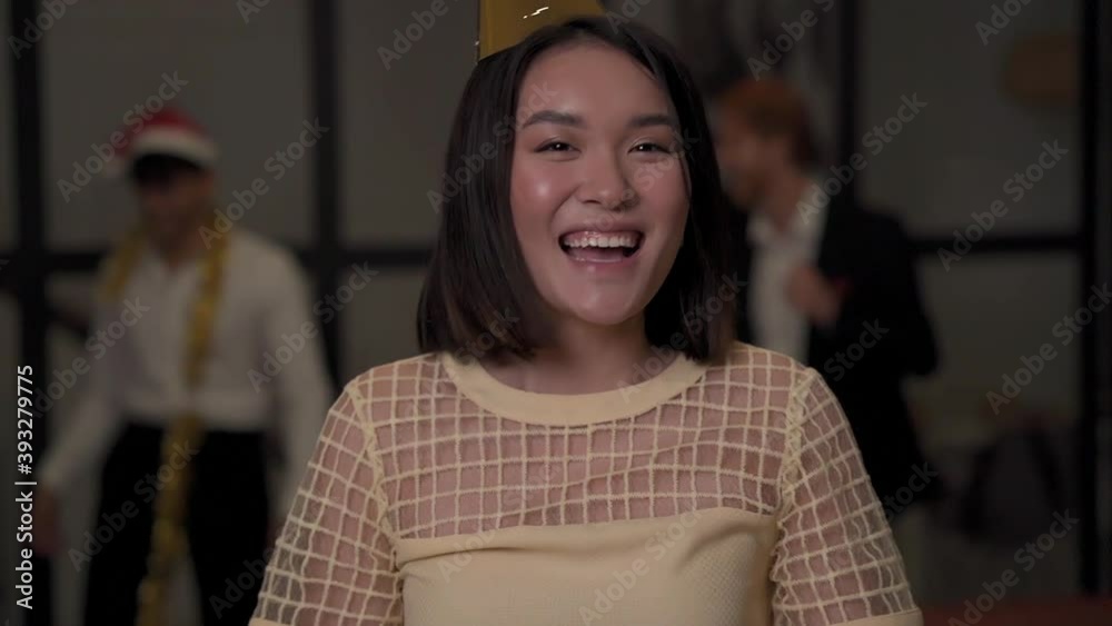 Portrait of young pretty asian girl blowing glitter confetti and looking at camera at New Year's Eve party. Christmas at home. Front view.