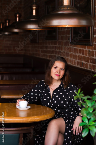Portrait of beautiful woman drinking a cup of coffee while sitting near the window in modern loft cafe bar. Portrait of smiling lady looking at camera.