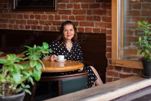 Portrait of beautiful woman drinking a cup of coffee while sitting near the window in modern loft cafe bar. Portrait of smiling lady looking at camera.