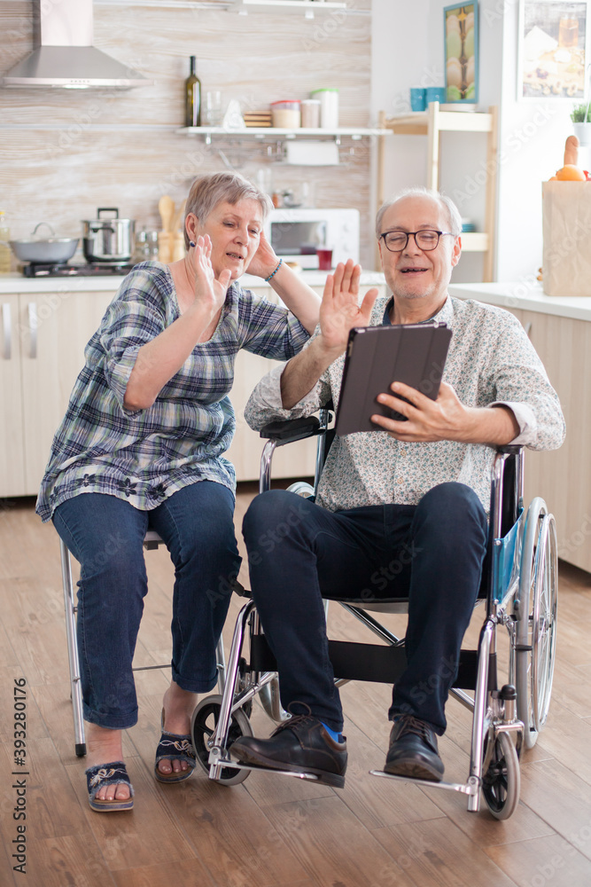Cheerful senior woman waving on video conference in the kitchen. Disabled senior man in wheelchair and his wife having a video conference on tablet pc in kitchen. Paralyzed old man and his wife having Cheerful senior woman waving on video conference in the kitchen. Disabled senior man in wheelchair and his wife having a video conference on tablet pc in kitchen. Paralyzed old man and his wife having