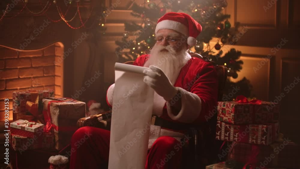 Santa Claus is reading list of good kids and checking gifts for Christmas, sitting in his decorated room at festive evening