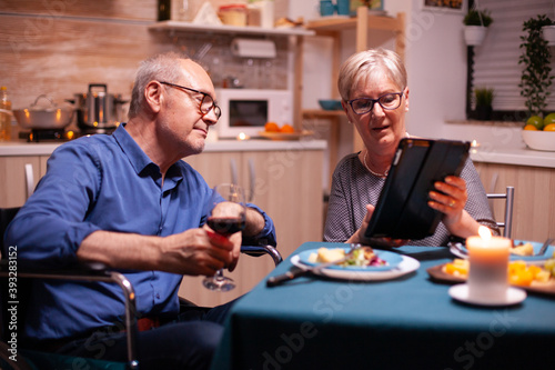 Elderly woman browsing on tablet pc and disabled husband in wheelchair holding wine glass. Imobilized handicapped senior husband browsing on phone enjoying the festive male  drinking a glass of red