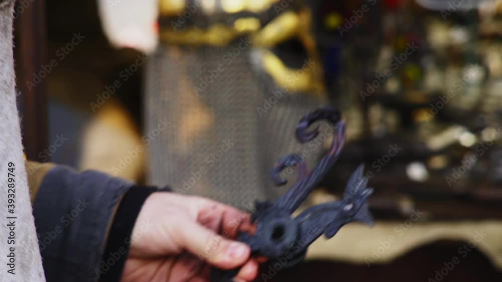 experienced smith in brown coat performs work making grey metal weather vane in cock form and holding in hands on famous Easter market on street of Prague