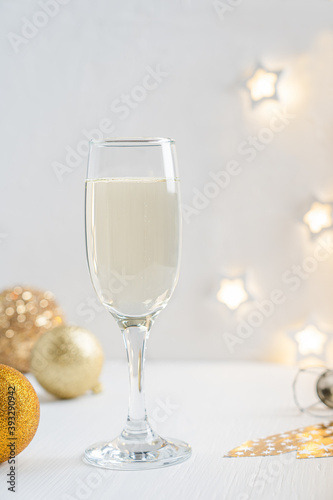 Cold white Prosecco sparkling champagne served in tall drinking glass with christmas or new year golden ball decoration and defocus garland lights on white wooden background. Vertical image