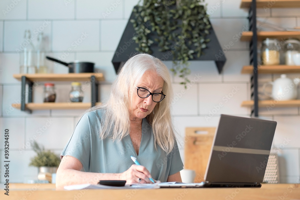 Senior woman using laptop for websurfing in her kitchen. The concept of senior employment, social security. Mature lady sitting at work typing a notebook computer in an home office. Senior woman using laptop for websurfing in her kitchen. The concept of senior employment, social security. Mature lady sitting at work typing a notebook computer in an home office.