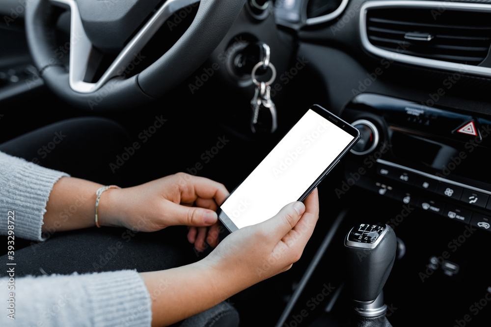 Obraz premium Young driver using smartphon in the car while driving or parking. Mockup of a mobile phone with isolated white screen in the hands of a woman.