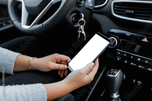 Young driver using smartphon in the car while driving or parking. Mockup of a mobile phone with isolated white screen in the hands of a woman.