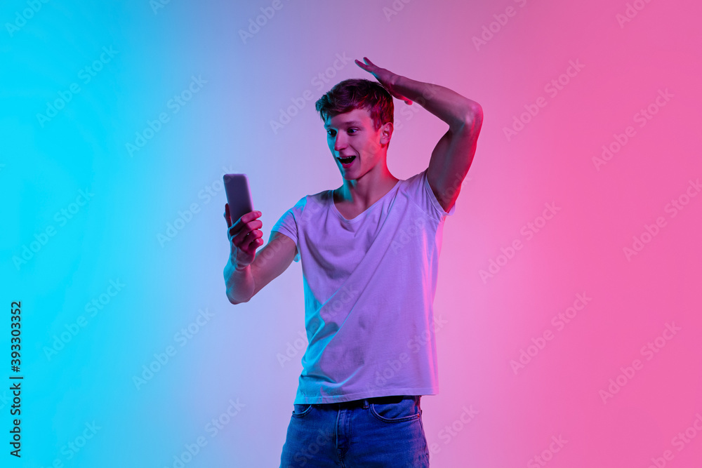 Shocked with cellphone. Young caucasian man's portrait on gradient blue-pink studio background in neon light. Concept of youth, human emotions, facial expression, sales, ad. Beautiful model in casual. Shocked with cellphone. Young caucasian man's portrait on gradient blue-pink studio background in neon light. Concept of youth, human emotions, facial expression, sales, ad. Beautiful model in casual.