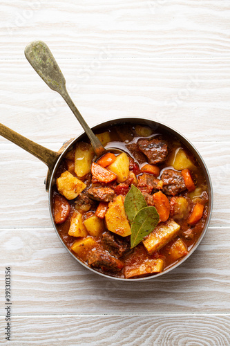 Delicious stew with meat, potatoes, carrot, herbs and gravy in rustic copper pot on white wooden background from above. Traditional winter and autumn dish beef and vegetables ragout in stewpot